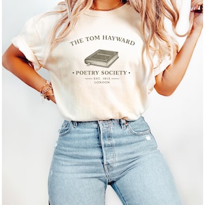 Puede incluir: Una camiseta de manga corta de color crema con un gráfico oscuro de un libro y el texto THE TOM HAYWARD POETRY SOCIETY EST. 1813 LONDON. La camiseta se combina con unos vaqueros de mezclilla azul claro con detalles desgastados en la parte delantera.