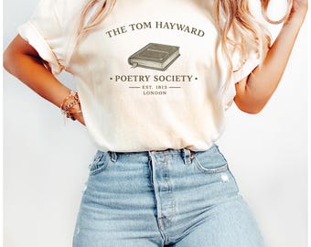 以下が含まれることがあります： クリーム色の半袖Tシャツで、本が描かれたダークなグラフィックと「THE TOM HAYWARD POETRY SOCIETY EST. 1813 LONDON」という文字がプリントされています。Tシャツには、フロントにダメージ加工が施されたライトブルーのデニムジーンズを合わせています。