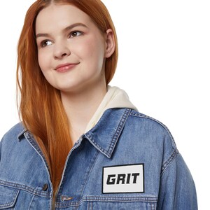 Könnte beinhalten: Hellblaue Jeansjacke mit einem weißen Aufnäher mit dem Schriftzug "GRIT" in schwarzen Buchstaben. Die Jacke hat ein klassisches Design mit Knopfverschlüssen und Taschen. Ein cremefarbener Kapuzenpullover wird darunter getragen.
