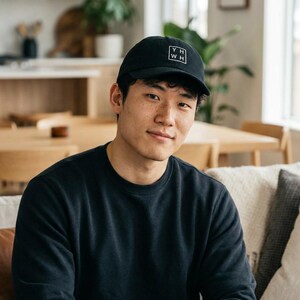 Può includere: Cappellino da baseball nero con un logo quadrato bianco con le lettere "YH" sopra "WH". Il cappellino è indossato da una persona che indossa una felpa nera. La persona è seduta su un divano.