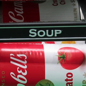 May include: A display of canned soup, featuring a red and white Campbell's Tomato Soup can with a large tomato illustration. The word "SOUP" is displayed above the cans in white letters on a black background.