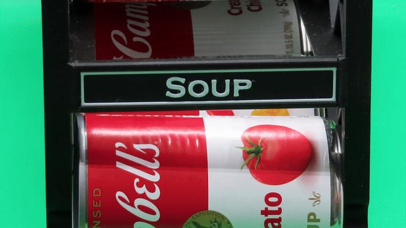 May include: A display of canned soup, featuring a red and white Campbell's Tomato Soup can with a large tomato illustration. The word "SOUP" is displayed above the cans in white letters on a black background.