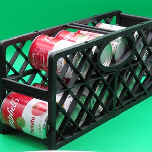 May include: A black plastic storage crate filled with several cans of Campbell's tomato soup and other canned goods. The crate has a diamond-shaped lattice design and is set against a green background.