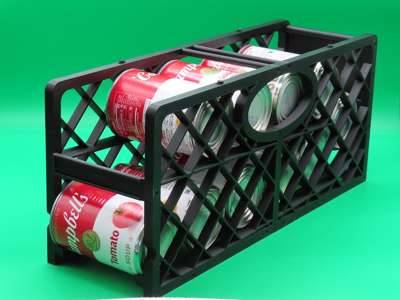 May include: A black plastic storage crate filled with several cans of Campbell's tomato soup and other canned goods. The crate has a diamond-shaped lattice design and is set against a green background.
