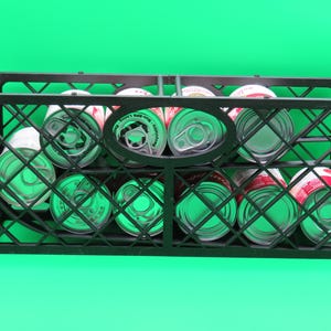 May include: A black plastic crate filled with canned goods, set against a bright green background. The cans are visible through the crate's grid-like structure, showcasing their red and white labels and silver tops.