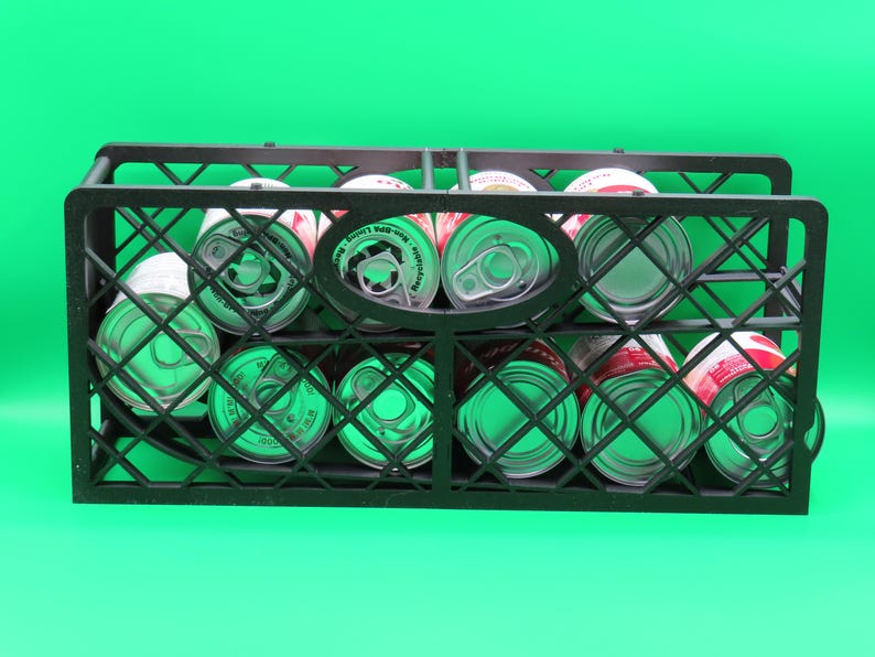 May include: A black plastic crate filled with canned goods, set against a bright green background. The cans are visible through the crate's grid-like structure, showcasing their red and white labels and silver tops.