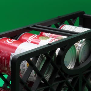 May include: A black plastic crate filled with several cans of soup. The cans are red and white with the brand name visible. The crate has a geometric design and is set against a green background.