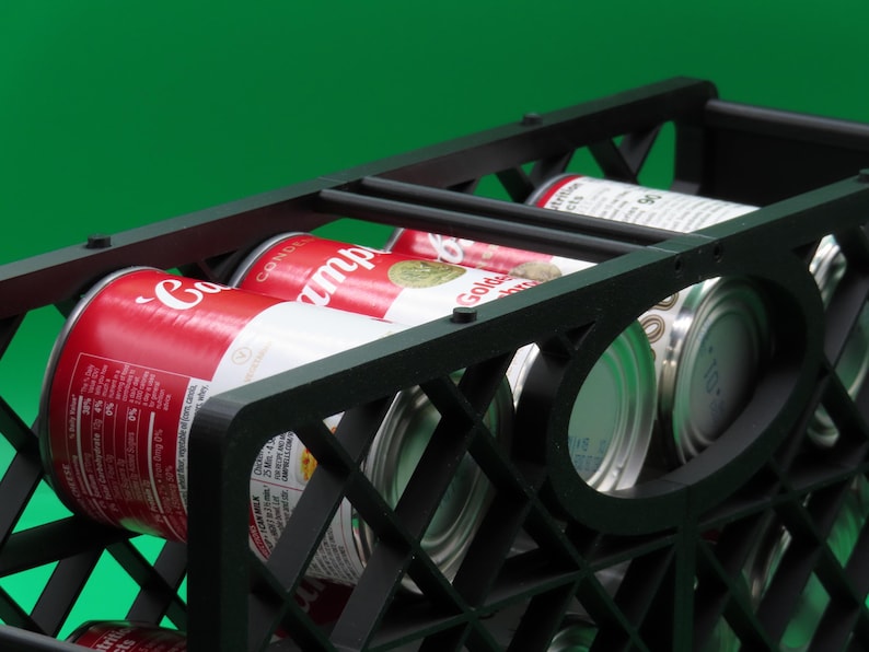 May include: A black plastic crate filled with several cans of soup. The cans are red and white with the brand name visible. The crate has a geometric design and is set against a green background.