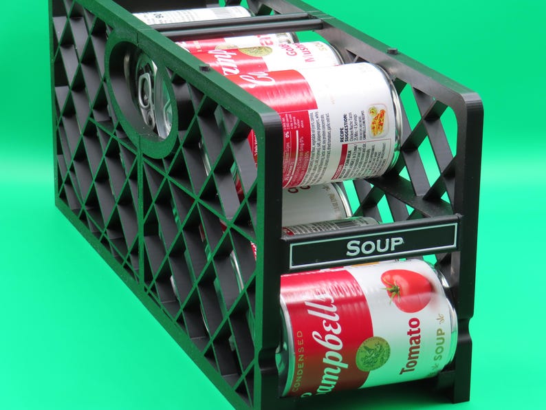 May include: Black plastic can organizer holding several cans of Campbell's soup. The cans are red and white with the Campbell's logo and tomato graphics. The organizer has a diamond-patterned design and a label that reads "SOUP."
