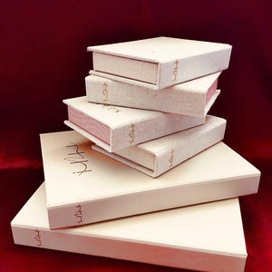 Può includere: Una pila di scatole bianco sporco, con texture di lino e scritte dorate. Le scatole sono rettangolari e simili a libri, alcune con bordi a righe rosse. Sono disposte su una superficie rosso scuro.