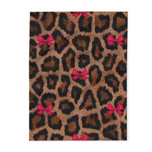 Puede incluir: Una manta rectangular con un estampado de leopardo en tonos marrones y negros. Hay lazos rosas brillantes distribuidos por toda la superficie. La manta parece estar hecha de un material suave.