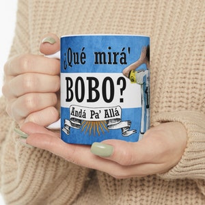 May include: A white ceramic mug with a blue and white design featuring a soccer player and the text "&iquest;Qu&eacute; mir&aacute;' Bobo? And&aacute; Pa' All&aacute;".