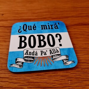 May include: Blue and white magnet with the text "¿Qué mirá' Bobo? Andá Pa' Allá" in Spanish. The magnet features a stylized sun and a ribbon with a scroll.