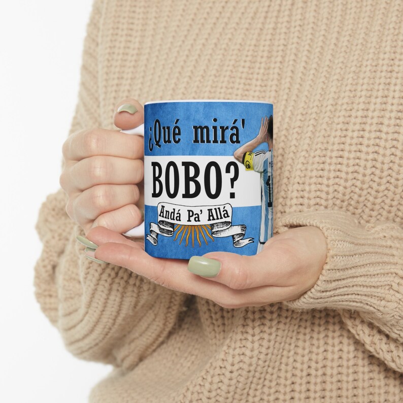 May include: A white ceramic mug with a blue and white design featuring a soccer player and the text "&iquest;Qu&eacute; mir&aacute;' Bobo? And&aacute; Pa' All&aacute;".