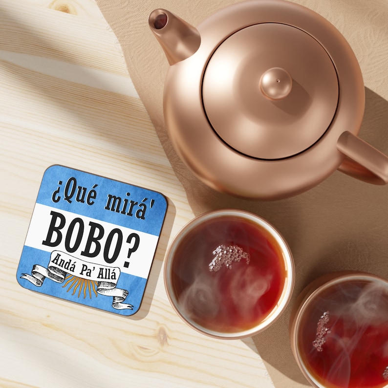 May include: A blue and white coaster with the text "&iquest;Qu&eacute; mir&aacute;' Bobo? And&aacute; Pa' All&aacute;" in black. The coaster is sitting on a wooden surface next to a copper teapot and two cups of tea.