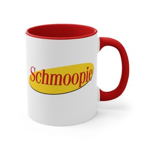 May include: White ceramic mug with a red rim and handle. The mug features a yellow oval with the word "Schmoopie" in red letters.
