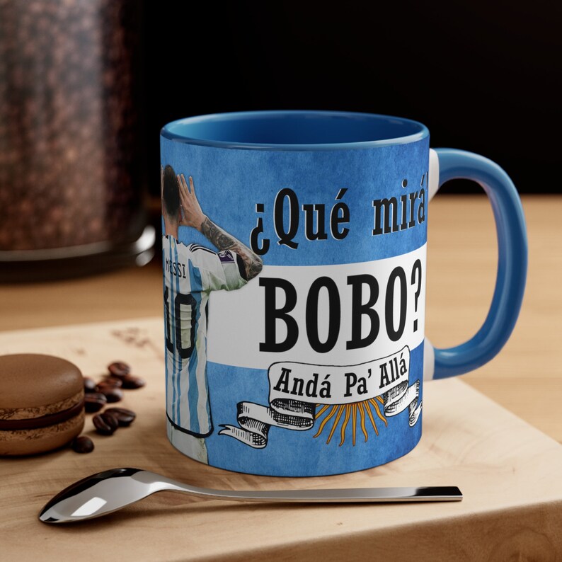 May include: A blue ceramic mug with a white and blue design. The mug features a soccer player in a blue and white jersey, the text "&iquest;Qu&eacute; mira Bobo? And&aacute; Pa' All&aacute;", and a sun with rays.