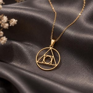 May include: A gold-coloured necklace featuring a circular pendant with a geometric design. The pendant consists of an outer circle enclosing a triangle, which contains a smaller central circle. The piece hangs from a delicate gold-coloured chain.