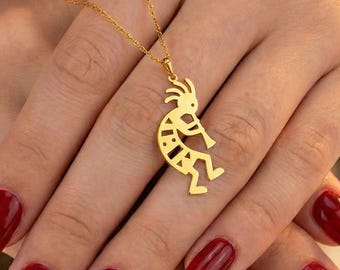 Pode incluir: Um colar dourado com um pingente Kokopelli, retratando um flautista. O pingente pende de uma corrente fina. O fundo mostra uma mão com verniz de unhas vermelho.
