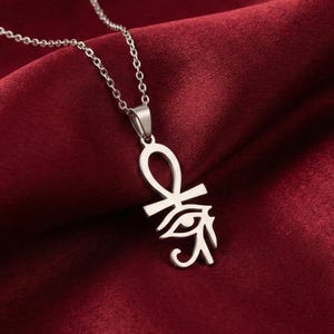 May include: A silver-colored necklace with a pendant featuring an Ankh symbol and the Eye of Horus. The necklace hangs against a deep red satin fabric, creating a striking contrast. The chain is delicate and the pendant is detailed.