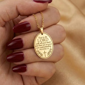 Op de afbeelding: Een goudkleurige ovale hanger aan een ketting met een gegraveerde tekst die luidt Trust in the LORD with all your heart and lean not on your own understanding. PROVERBS 3:5, voorzien van een klein kruisje en bloemmotieven.