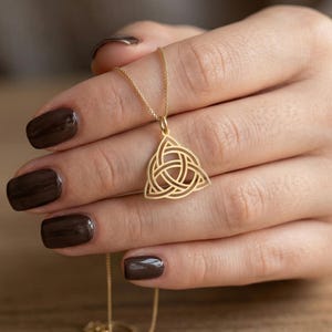 Könnte beinhalten: Eine goldfarbene Halskette mit einem keltischen Knotenanhänger. Der Anhänger hat ein detailliertes, offenes Design in Form eines Dreiecks. Die Halskette wird vor einem verschwommenen Hintergrund gehalten, der die filigrane Handwerkskunst zeigt.