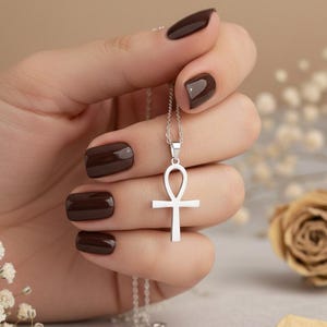 May include: A silver ankh pendant necklace featuring a polished metallic finish. The pendant is shaped like an Egyptian ankh cross with a loop at the top and a vertical bar extending downward, suspended from a delicate silver chain link necklace.
