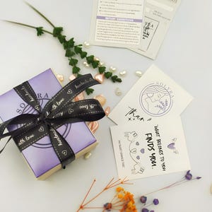K&ouml;nnte beinhalten: Eine kleine, lavendelfarbene Geschenkbox, gebunden mit einem schwarzen Band mit der Aufschrift "handmade with love". Die Box ist von dekorativen Elementen umgeben, darunter Karten mit den Texten "SOLYRA" und "What belongs to you finds you."