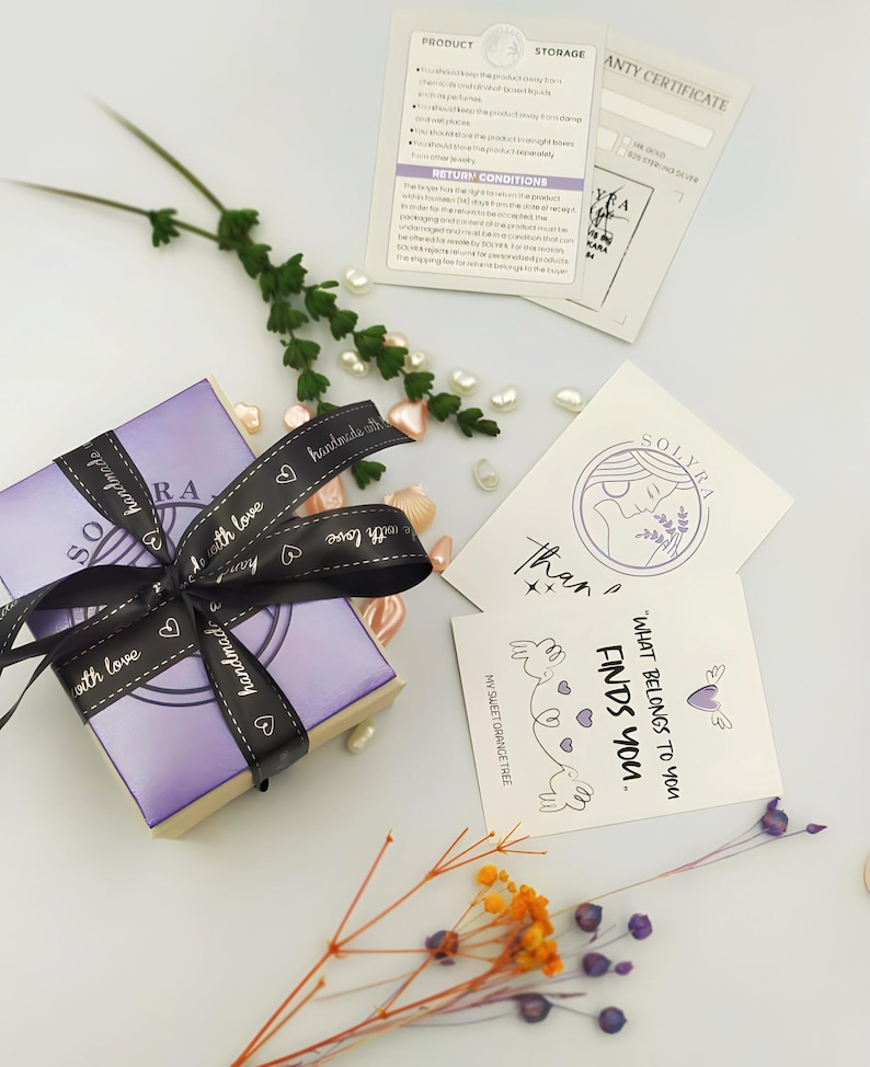 K&ouml;nnte beinhalten: Eine kleine, lavendelfarbene Geschenkbox, gebunden mit einem schwarzen Band mit der Aufschrift "handmade with love". Die Box ist von dekorativen Elementen umgeben, darunter Karten mit den Texten "SOLYRA" und "What belongs to you finds you."