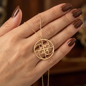 May include: A gold pendant necklace with a geometric design. The pendant is circular with an intricate pattern of lines and curves. The necklace is held in a hand with brown nail polish.