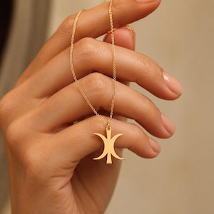 May include: A gold-toned necklace featuring a delicate chain and a central pendant shaped like a stylized triple crescent moon symbol. The pendant has a smooth, polished finish and is suspended from a thin, matching gold-colored metal chain.