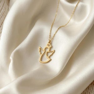 K&ouml;nnte beinhalten: Eine goldfarbene Engels-Anh&auml;nger-Halskette. Der Anh&auml;nger zeigt eine Engels-Silhouette mit ausgebreiteten Fl&uuml;geln und einem Heiligenschein. Die Halskette hat eine feine goldene Kette und liegt auf wei&szlig;em Satin.