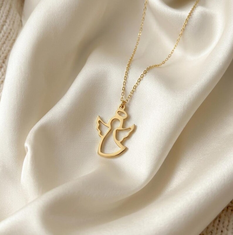 K&ouml;nnte beinhalten: Eine goldfarbene Engels-Anh&auml;nger-Halskette. Der Anh&auml;nger zeigt eine Engels-Silhouette mit ausgebreiteten Fl&uuml;geln und einem Heiligenschein. Die Halskette hat eine feine goldene Kette und liegt auf wei&szlig;em Satin.