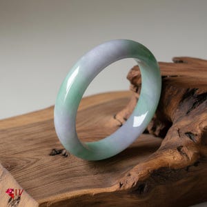 May include: A smooth, polished bangle bracelet crafted from translucent jade stone. The circular accessory features a gradient color palette transitioning between soft lavender and pale green hues, with a glossy finish that reflects light.
