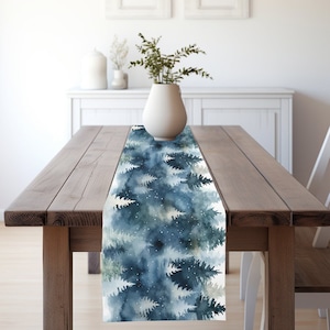 May include: A blue and white watercolor table runner with a forest scene. The runner features a repeating pattern of pine trees and snowflakes.