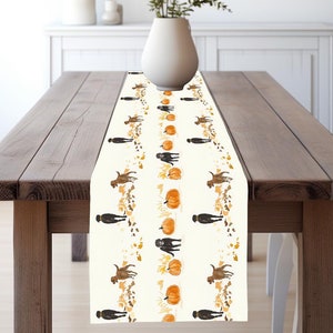 May include: A white table runner with a repeating pattern of brown dogs, orange pumpkins, and gold leaves. The runner is on a wooden table with a vase of green plants.
