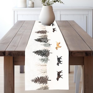 May include: White table runner with a winter forest scene featuring brown and gray trees with snow and black and brown dogs walking through the trees.