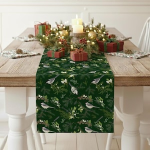 Peut inclure: Une table à manger en bois dressée pour un repas de fête. Un chemin de table vert avec un motif d'oiseaux et de plantes orne le centre. La table est décorée de cadeaux, de bougies et de verdure. Des chaises blanches entourent la table.