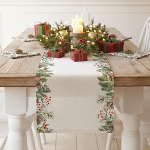May include: A festive Christmas table setting with a wooden table, white chairs, and a cream-colored table runner. The runner is decorated with green pine branches, red berries, and pine cones. Small red gift boxes and candles complete the holiday scene.