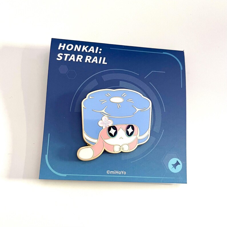 May include: A pin featuring a cartoon character with a blue and white design, with a pink and white character in front of a blue object. The pin is on a blue card with the text "HONKAI: STAR RAIL". The copyright reads "&copy;miHoYo".