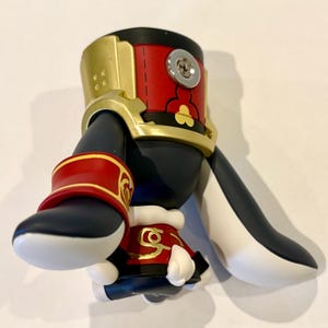 May include: A stylized figurine featuring a black and white body with red and gold accents. The figure has a red cylindrical top section with a silver screw, a gold collar, and red arm bands with gold trim. The base is black with white details.