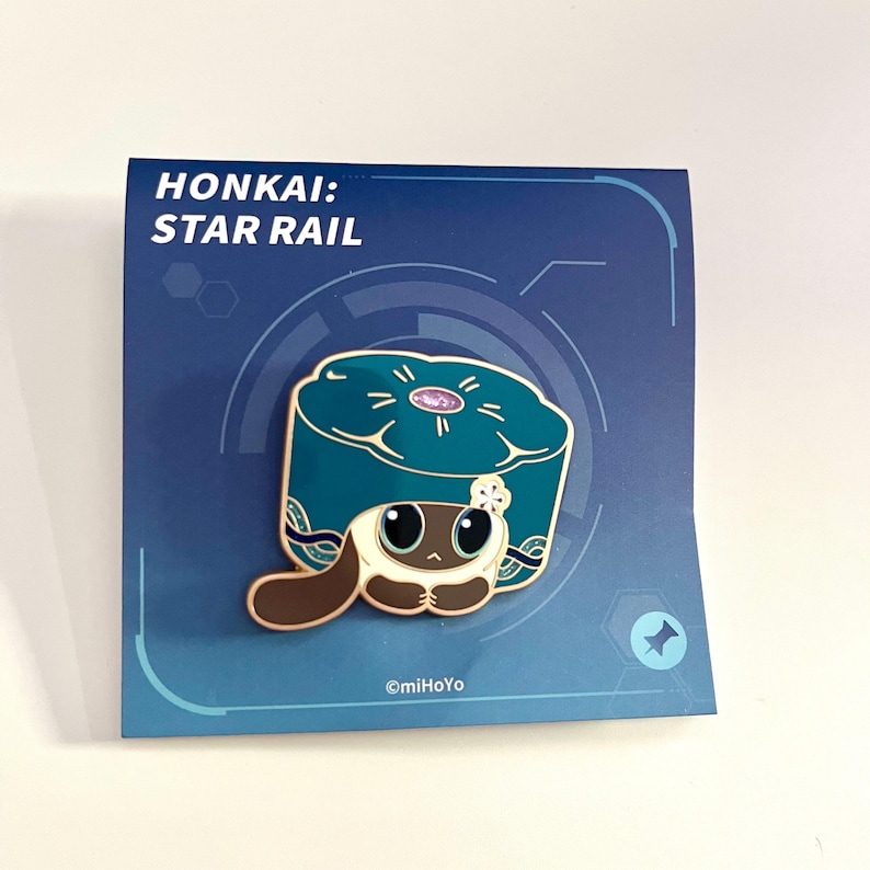 May include: Enamel pin featuring a cartoon character with large eyes, brown and white fur, and a teal hat. The pin is on a blue card with the text "HONKAI: STAR RAIL". The pin has gold-colored outlines and a flower detail.