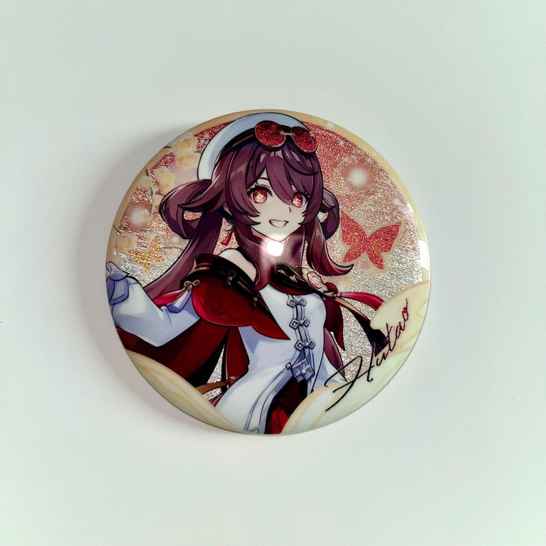 Pu&ograve; includere: Bottone rotondo con un'illustrazione di un personaggio anime sorridente con lunghi capelli castano scuro, che indossa un abito bianco e rosso. Lo sfondo presenta glitter e farfalle, con il nome "Hutao" in corsivo.