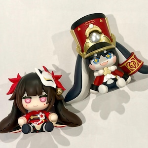 May include: Two stylized chibi-style figurines. One figure has dark hair, a white fox mask, and a red outfit. The second figure wears a tall red and gold military-style hat, a red jacket with white accents, and has long dark ears extending downwards.