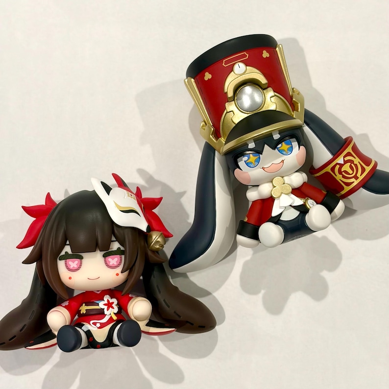 May include: Two stylized chibi-style figurines. One figure has dark hair, a white fox mask, and a red outfit. The second figure wears a tall red and gold military-style hat, a red jacket with white accents, and has long dark ears extending downwards.