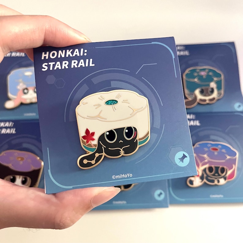 May include: A pin featuring a black cartoon character with large eyes, wearing a white hat with a red flower. The pin is on a blue card with the text "HONKAI: STAR RAIL". Other pins are visible in the background.