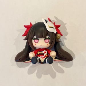 May include: A small chibi-style figurine with dark brown hair, wearing a red outfit with a white flower accent. The figure has pink eyes with cross patterns, a white fox mask perched on the head, and red hair accessories with a small gold bell.