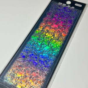 Può includere: Una confezione rettangolare contenente fogli di adesivi olografici con una varietà di piccole forme, tra cui stelle, fiocchi di neve e cerchi. Gli adesivi presentano una finitura riflettente color arcobaleno che cambia a seconda della luce.