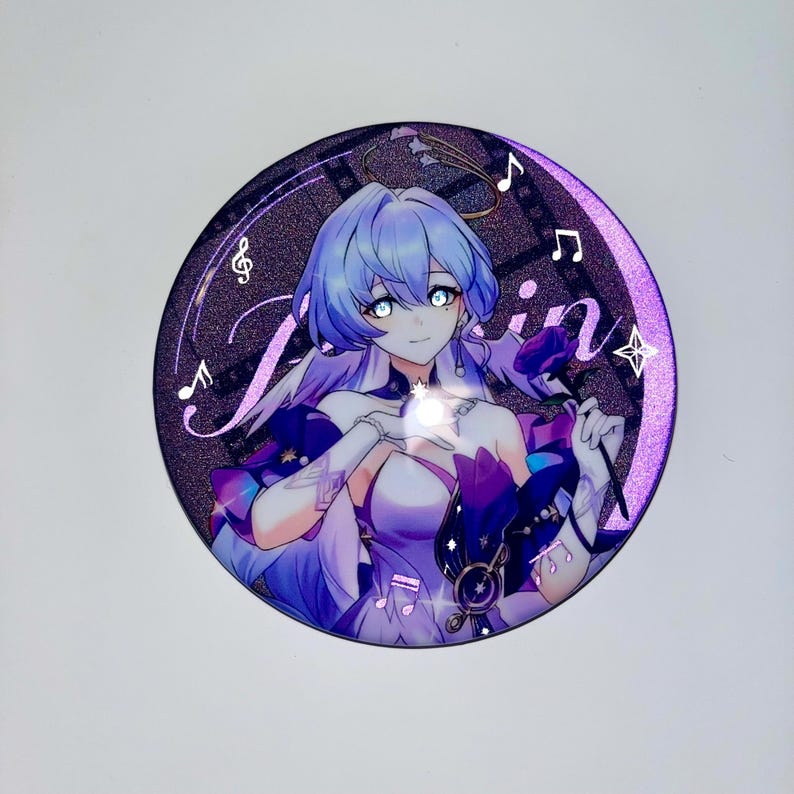 May include: A round, purple and silver glitter sticker featuring an anime character with long, blue hair. The character is wearing a white and purple dress and holding a rose. The sticker has a film strip design and musical notes around the edge. The text "Ganyu" is written in a stylized font on the sticker.