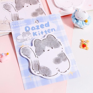Puede incluir: Un paquete de notas adhesivas "Dazed Kitten" con un dise&ntilde;o de gato de dibujos animados en gris y blanco. El paquete est&aacute; sobre un fondo a cuadros azul, con otros elementos decorativos, incluido un peque&ntilde;o oso con una fresa.